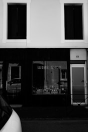 A black and white photo of a storefront labeled 'Wine Shop'. The shop has a window display featuring several wine bottles. A parked vehicle is partially visible in the foreground, and the building facade includes two rectangular windows above the shop.