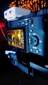 A digital camera with a screen displaying a live view of people gathered under warm lighting. The camera's various buttons and settings are visible, with a focus on the display screen showing the image composition.