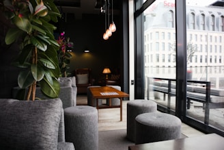 A modern, cozy lounge area featuring a wooden table surrounded by grey upholstered seating and stools. Large glass windows reveal a view of a contemporary building exterior. The lighting is warm with hanging pendant lights and a table lamp providing a relaxed atmosphere. Plants add a touch of greenery, and the overall decor is minimalist yet inviting.