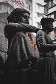 A dark bronze statue features a seated figure adorned with intricately detailed clothing and garlands of orange and white flowers. A second figure stands nearby. Buildings and large text in another language form the background.