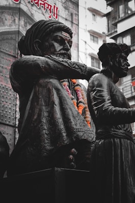 A dark bronze statue features a seated figure adorned with intricately detailed clothing and garlands of orange and white flowers. A second figure stands nearby. Buildings and large text in another language form the background.