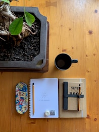 A wooden table with a potted plant with lush green leaves on the left. Next to the plant, there is a black coffee mug. Below, there's a colorful pencil case with comic-style patterns, an open spiral notebook with a pencil, an eraser, and a sharpener on it, with 'Yo' and 'Lo m&aacute;s importante' written in Spanish. Next to the notebook, there are two closed notebooks, one with a cover that says 'Never judge a book by its cover' and another identical pencil on top.