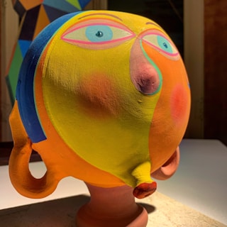 A colorful, abstract sculpture resembling a face with exaggerated features, painted with vibrant shades of yellow, orange, blue, and pink. The background features a geometric abstract painting with similar colors.