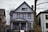 A large, aged house with a weathered purple facade stands between two other homes. The structure has multiple windows, a prominent gable roof, and a porch. Bare trees surround it, and there is a wheelchair ramp leading to the door. The house appears worn, with visible signs of peeling paint and damage.