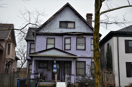 A large, aged house with a weathered purple facade stands between two other homes. The structure has multiple windows, a prominent gable roof, and a porch. Bare trees surround it, and there is a wheelchair ramp leading to the door. The house appears worn, with visible signs of peeling paint and damage.