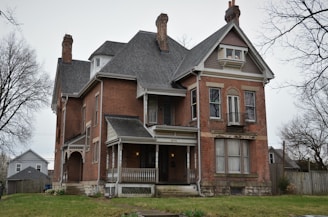 A large, historic brick house with two prominent chimneys and a pitched roof. The house features multiple windows, some with decorative trim and shutters, and a small covered porch with columns at the front entrance. The lawns are bordered by a wooden fence, and leafless trees are visible in the background under an overcast sky.