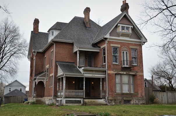 A large, historic brick house with two prominent chimneys and a pitched roof. The house features multiple windows, some with decorative trim and shutters, and a small covered porch with columns at the front entrance. The lawns are bordered by a wooden fence, and leafless trees are visible in the background under an overcast sky.