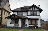 A two-story house with a Tudor-style architectural design featuring dark timber frames against white walls. The house has a prominent front porch supported by wooden beams. There are two visible windows on the upper floor and a landscaped front yard with shrubs. Adjacent to this house is another building with different architectural details.