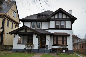 A two-story house with a Tudor-style architectural design featuring dark timber frames against white walls. The house has a prominent front porch supported by wooden beams. There are two visible windows on the upper floor and a landscaped front yard with shrubs. Adjacent to this house is another building with different architectural details.