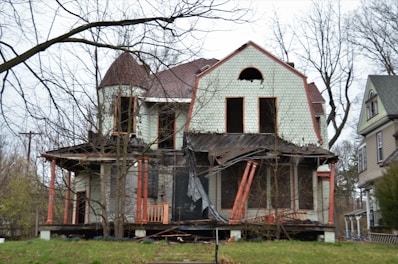 A dilapidated, abandoned house with visible structural damage, including a partially collapsed porch and broken windows. The siding is peeling, and the roof shows signs of disrepair. Bare trees surround the house, emphasizing the neglected state.