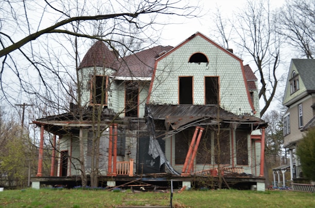 A dilapidated, abandoned house with visible structural damage, including a partially collapsed porch and broken windows. The siding is peeling, and the roof shows signs of disrepair. Bare trees surround the house, emphasizing the neglected state.