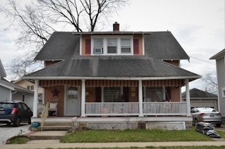 A two-story house with a large front porch. The house has a steeply pitched roof with dark shingles and features two dormer windows on the second floor. The porch is covered and supported by white columns. There is a star decoration on the wall next to the front door. Several items such as chairs and plants are visible on the porch. A tree with bare branches stands behind the house. On either side of the house, there are neighboring houses and parked cars.