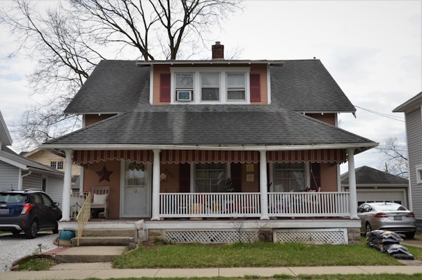 A two-story house with a large front porch. The house has a steeply pitched roof with dark shingles and features two dormer windows on the second floor. The porch is covered and supported by white columns. There is a star decoration on the wall next to the front door. Several items such as chairs and plants are visible on the porch. A tree with bare branches stands behind the house. On either side of the house, there are neighboring houses and parked cars.