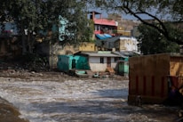 Several buildings in disrepair are close to a turbulent water body. The buildings appear weathered and some are dilapidated. There is debris scattered around the buildings. Tall trees and some vegetation are present near the structures. The sky is clear with bright daylight.