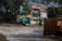 Several buildings in disrepair are close to a turbulent water body. The buildings appear weathered and some are dilapidated. There is debris scattered around the buildings. Tall trees and some vegetation are present near the structures. The sky is clear with bright daylight.