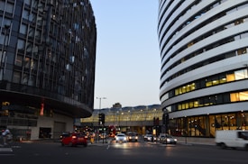 An urban street scene featuring two modern buildings with unique architectural designs on either side. The left building has a dark, reflective facade with an irregular pattern, while the right building has white bands curving around it. In the background, a glass and metal structure bridges the space between the buildings. Several vehicles, including cars and buses, are visible on the street, and there are people walking and cycling.