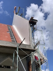 A building under construction with visible scaffolding and a worker wearing a red shirt on the platform. The structure has a red tiled roof and sections of exposed concrete and bricks. The sky is clear with a few clouds.
