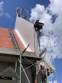 A building under construction with visible scaffolding and a worker wearing a red shirt on the platform. The structure has a red tiled roof and sections of exposed concrete and bricks. The sky is clear with a few clouds.