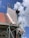 A building under construction with visible scaffolding and a worker wearing a red shirt on the platform. The structure has a red tiled roof and sections of exposed concrete and bricks. The sky is clear with a few clouds.