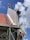 A building under construction with visible scaffolding and a worker wearing a red shirt on the platform. The structure has a red tiled roof and sections of exposed concrete and bricks. The sky is clear with a few clouds.