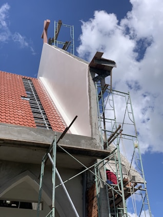 A building under construction with visible scaffolding and a worker wearing a red shirt on the platform. The structure has a red tiled roof and sections of exposed concrete and bricks. The sky is clear with a few clouds.