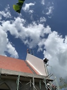 A building under construction with a partially completed roof featuring red tiles and scaffolding on its side. The sky is filled with fluffy white clouds against a backdrop of blue. A large green leaf appears in the top left corner.