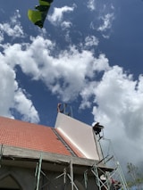 A building under construction with a partially completed roof featuring red tiles and scaffolding on its side. The sky is filled with fluffy white clouds against a backdrop of blue. A large green leaf appears in the top left corner.
