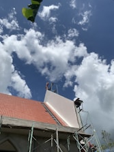 A building under construction with a partially completed roof featuring red tiles and scaffolding on its side. The sky is filled with fluffy white clouds against a backdrop of blue. A large green leaf appears in the top left corner.