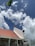 A building under construction with a partially completed roof featuring red tiles and scaffolding on its side. The sky is filled with fluffy white clouds against a backdrop of blue. A large green leaf appears in the top left corner.