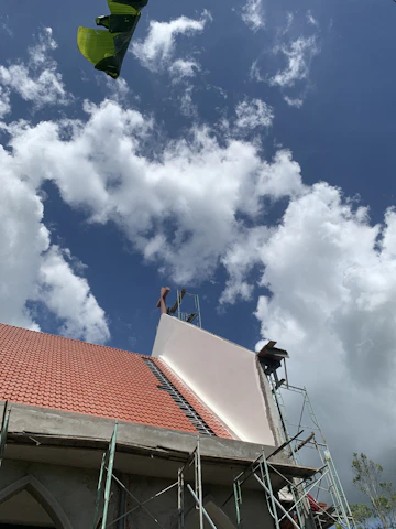 A building under construction with a partially completed roof featuring red tiles and scaffolding on its side. The sky is filled with fluffy white clouds against a backdrop of blue. A large green leaf appears in the top left corner.