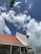A building under construction with a partially completed roof featuring red tiles and scaffolding on its side. The sky is filled with fluffy white clouds against a backdrop of blue. A large green leaf appears in the top left corner.