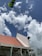 A building under construction with a partially completed roof featuring red tiles and scaffolding on its side. The sky is filled with fluffy white clouds against a backdrop of blue. A large green leaf appears in the top left corner.