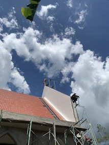 A building under construction with a partially completed roof featuring red tiles and scaffolding on its side. The sky is filled with fluffy white clouds against a backdrop of blue. A large green leaf appears in the top left corner.