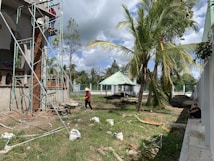 A construction site with scaffolding set up around a building partially made of bricks and concrete. Two workers are present; one is on the scaffolding, and the other is walking on the ground wearing protective gear. The area is surrounded by palm trees, and there is a small pavilion with a green roof in the background. Various construction materials and debris are scattered on the grass.