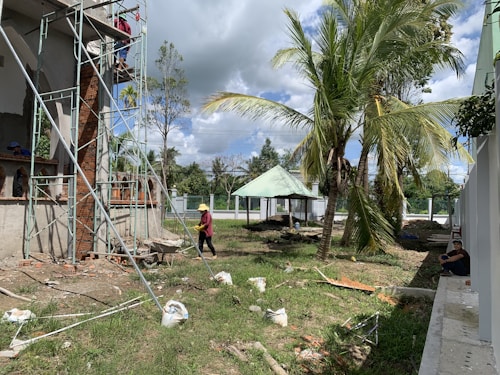A construction site with scaffolding set up around a building partially made of bricks and concrete. Two workers are present; one is on the scaffolding, and the other is walking on the ground wearing protective gear. The area is surrounded by palm trees, and there is a small pavilion with a green roof in the background. Various construction materials and debris are scattered on the grass.