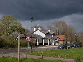 A quaint pub is situated by the side of a road, bordered by lush greenery and tall trees. The building features traditional architecture with white walls, dark timber frames, and a red-tiled roof. Hanging flower baskets add a touch of color to the exterior. Several cars are parked along the road, and a lamp post stands prominently nearby. The sky is overcast with dramatic dark clouds, casting a shadow over the scene.