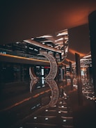 A dimly lit shopping mall interior featuring modern architecture and a large decorative crescent structure made of sparkling lights. The floor is shiny, reflecting the lights and the structure, creating an elegant ambiance. Various retail stores are visible along the sides with illuminated signage.