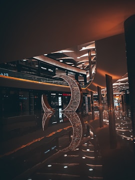 A dimly lit shopping mall interior featuring modern architecture and a large decorative crescent structure made of sparkling lights. The floor is shiny, reflecting the lights and the structure, creating an elegant ambiance. Various retail stores are visible along the sides with illuminated signage.