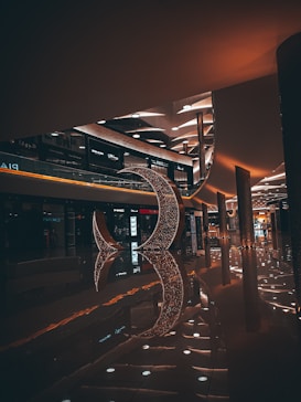 A dimly lit shopping mall interior featuring modern architecture and a large decorative crescent structure made of sparkling lights. The floor is shiny, reflecting the lights and the structure, creating an elegant ambiance. Various retail stores are visible along the sides with illuminated signage.