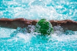 A swimmer is performing the butterfly stroke in a pool. Their muscular arms are outstretched, with splashes of water around them. The swimmer is wearing a green swim cap with white text, submerged partially in the clear, blue water.