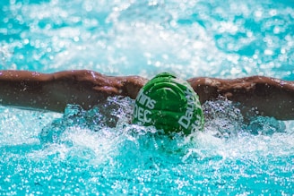 A swimmer is performing the butterfly stroke in a pool. Their muscular arms are outstretched, with splashes of water around them. The swimmer is wearing a green swim cap with white text, submerged partially in the clear, blue water.
