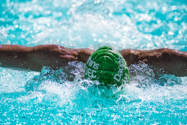 A swimmer is performing the butterfly stroke in a pool. Their muscular arms are outstretched, with splashes of water around them. The swimmer is wearing a green swim cap with white text, submerged partially in the clear, blue water.