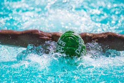 A swimmer is performing the butterfly stroke in a pool. Their muscular arms are outstretched, with splashes of water around them. The swimmer is wearing a green swim cap with white text, submerged partially in the clear, blue water.