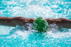 A swimmer is performing the butterfly stroke in a pool. Their muscular arms are outstretched, with splashes of water around them. The swimmer is wearing a green swim cap with white text, submerged partially in the clear, blue water.