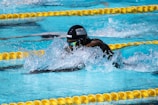 A swimmer is captured mid-stroke in a competitive swimming pool. Wearing a black swim cap with an American flag and reflective goggles, the swimmer is surrounded by splashing water. Yellow lane dividers line the pool, indicating lane boundaries.