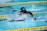 A swimmer is captured mid-stroke in a competitive swimming pool. Wearing a black swim cap with an American flag and reflective goggles, the swimmer is surrounded by splashing water. Yellow lane dividers line the pool, indicating lane boundaries.