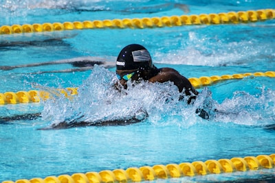 A swimmer is captured mid-stroke in a competitive swimming pool. Wearing a black swim cap with an American flag and reflective goggles, the swimmer is surrounded by splashing water. Yellow lane dividers line the pool, indicating lane boundaries.
