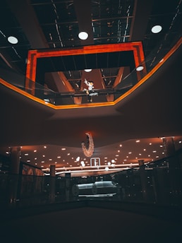 A modern interior space features a dramatic lighting arrangement with a warm orange glow framing a ceiling area. Numerous circular lights are embedded in the panels, illuminating a reflective surface. A distinctive chandelier hangs prominently in the center. An upper level walkway with glass railings is visible, along with decorative elements and signage.