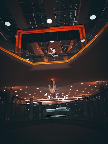 A modern interior space features a dramatic lighting arrangement with a warm orange glow framing a ceiling area. Numerous circular lights are embedded in the panels, illuminating a reflective surface. A distinctive chandelier hangs prominently in the center. An upper level walkway with glass railings is visible, along with decorative elements and signage.
