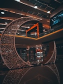 A crescent-shaped installation adorned with numerous small lights stands in a modern indoor space. The ambient lighting creates a warm and inviting atmosphere. The ceiling features contemporary design elements with recessed lighting and round fixtures. Reflections on the polished floor enhance the aesthetic appeal of the setting.